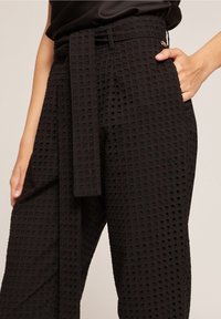 Pantalones negros con textura y estampado de cuadro, con una cinturilla ancha, trabillas para cinturón y un cinturón de tela atado. Incluye bolsillos laterales.