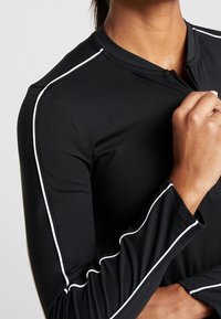Black long-sleeve top with a zip, featuring white accent piping along the shoulders and sleeves, made from a smooth, stretchy material.