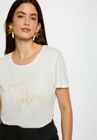 T-shirt bianco a maniche corte con testo in glitter oro che legge "Good Vibes". Tessuto morbido con scollatura rotonda, abbinato a pantaloni scuri.
