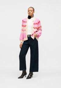 Γυναίκα που φοράει ροζ ζακέτα με σχέδια, λευκό ριμπ μπλουζάκι, σκουρόχρωμο τζιν παντελόνι με φαρδύ πόδι, μαύρες μπότες στον αστράγαλο και μαύρο κολιέ με λουλούδια, στέκεται με το ένα χέρι στην τσέπη.