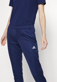 Námořnické modré športové nohavice s elastickým pásom, bočnými vreckami a bielym logom Adidas. Vyrobené z hladkej, ľahkej a priedušnej látky.