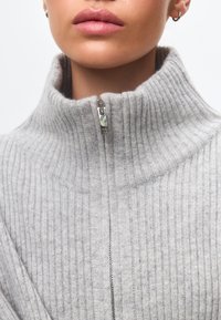 Grauer gerippter Pullover mit hohem Kragen und einem metallenen Reißverschluss am Hals. Der Stoff scheint weich mit einer strukturierten Oberfläche zu sein.