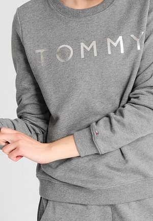 Henkilö, jolla on harmaa collegepaita, jossa on rinnan kohdalla "TOMMY"-teksti ja vasemman hiharesorin kohdalla pieni Tommy Hilfiger -logo.