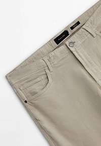 Pantalon beige en velours côtelé à coupe fuselée, avec poches avant, passants pour ceinture, fermeture par bouton métallique et étiquette de la marque Massimo Dutti visible à l’intérieur de la ceinture.