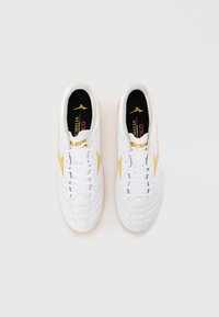 Hvite fotballsko med teksturert design, gule accenter og svarte innleggssåler med merker. Snøring langs fronten for sikker passform.