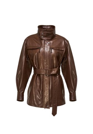 Nainen pukeutuneena vyölliseen ruskeaan nahkatakkiin, mustaan hameeseen ja mustiin polven yläpuolelle ulottuviin saappaisiin, käsissään musta käsilaukku, seisoo valkoista taustaa vasten.