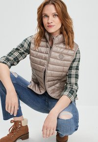Hellbraune Puffer-Weste mit hohem Kragen und Reißverschluss, kombiniert mit einem grün-weiß karierten Hemd, blauen, zerrissenen Jeans und braunen Schnürstiefeln.
