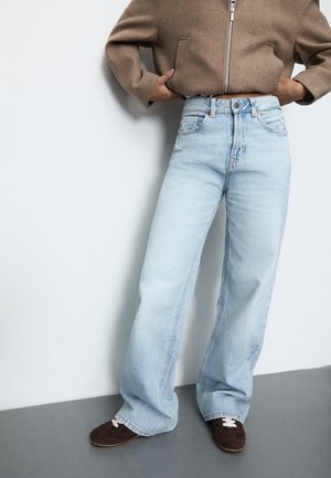 Personne portant un jean large bleu clair, une veste marron zippée et des chaussures marron foncé, debout avec les mains dans les poches de la veste.
