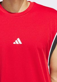 Crvena atletska majica bez rukava izrađena od teksturirane tkanine. Ima bijeli Adidas logo i kontrastne crno-bijele naglaske sa strane.