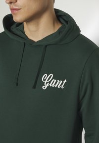 Homme portant un sweat à capuche vert foncé avec le texte blanc « Gant » sur la poitrine, montrant le cou et le bas du visage sur un fond neutre.