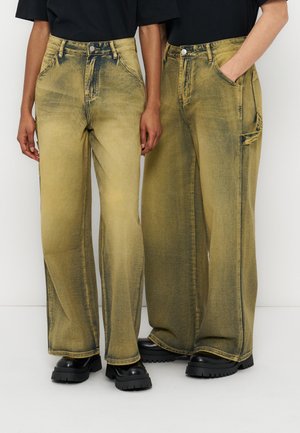 To personer står side om side iført falmede gule bukser med vide ben og sorte sko med kraftig sål, hænderne synlige, den ene hånd i lommen.