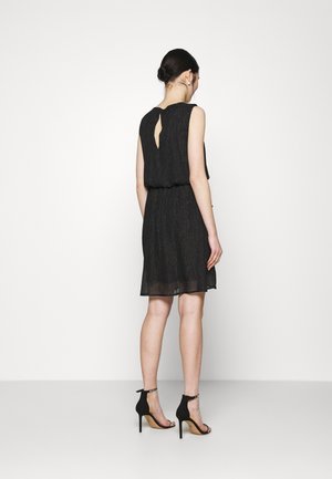 Robe noire en tissu léger et texturé. Présente un dos en forme de trou de serrure, une taille cintrée et une silhouette décontractée. Accompagnée de talons hauts noirs.