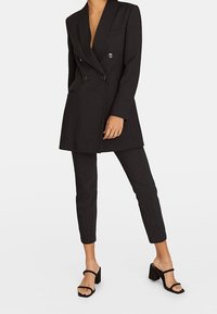 Vrouw in een zwarte dubbelgeknoopte blazer, bijpassende slim-fit cropped pants en zwarte hoge sandalen met enkelbandjes.