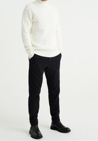 Muž v bílém texturovaném svetru, černých kordových kalhotách a černých kožených Chelsea botách, stojící s rukama v kapsách na bílém pozadí.