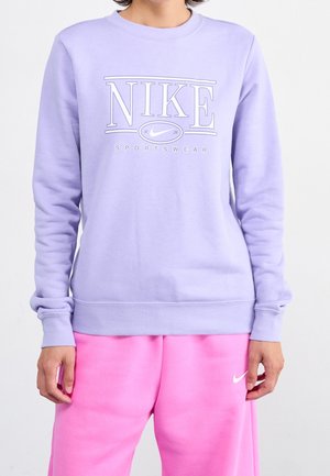 Persoon met een lavendelkleurige Nike-trui met wit logo en roze Nike-sweatpants tegen een effen lichte achtergrond.