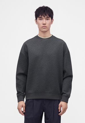 Junger Mann trägt ein dunkelgraues Sweatshirt mit Rundhalsausschnitt und dezentem Logo sowie dunkelblaue Hose und steht vor einem schlichten hellen Hintergrund.