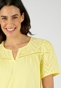 Geel blouse met korte mouwen, voorzien van ogen borduurwerk en een uitsnijding bij de hals. Zacht stof met een lichte textuur.