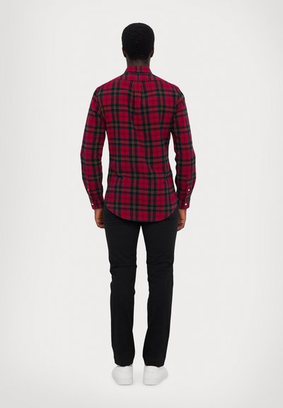 Κόκκινο καρό πουκάμισο με πράσινες και κίτρινες λεπτομέρειες, γιακάς με κουμπιά και καμπύλο στρίφωμα, συνδυασμένο με μαύρο παντελόνι και λευκά αθλητικά παπούτσια, φ viewed from the back.