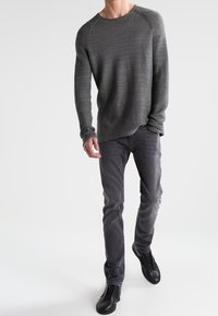 Homme portant un pull à manches longues gris texturé, un jean slim gris foncé et des chaussures noires sans lacets, debout devant un fond blanc.
