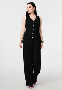 Veste noire sans manches avec un col en V et cinq boutons argentés, associée à un pantalon noir à jambes larges et des sandales pourpres ornées de plumes.