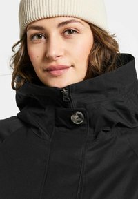 Zwarte weerbestendige jas met hoge kraag, ritssluiting en knopenaccent. Zachte textuur en minimalistisch ontwerp. Lichtbeige gebreide muts zichtbaar.