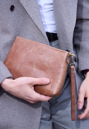 Beutelförmige Clutch aus cognacfarbenem Leder mit strukturierter Oberfläche, Reißverschluss und abnehmbarem Handgelenkriemen, in einer Hand gehalten vor einem grauen Mantel.