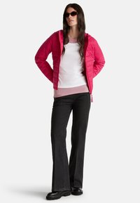 Žena s červenou prošívanou bundou přes bílé a růžové topy, černými zvonovými džínami, černými botami a tmavými slunečními brýlemi, stojí s rukama za zády.