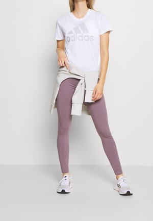 T-shirt Adidas blanche avec logo gris, associée à des leggings violet clair. Un sweat-shirt gris clair noué à la taille et des chaussures de sport blanches.