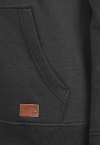 Sudadera negra con bolsillo tipo canguro, puños acanalados y dobladillo. Presenta una etiqueta de cuero marrón con el texto "BLEND GLOBAL JEANMAKERS".