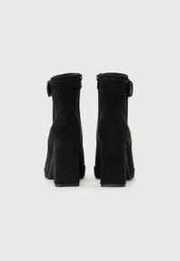 Par svart mocka ankelboots med klackar och ankelremmar, visade från baksidan på en enkel ljus bakgrund.