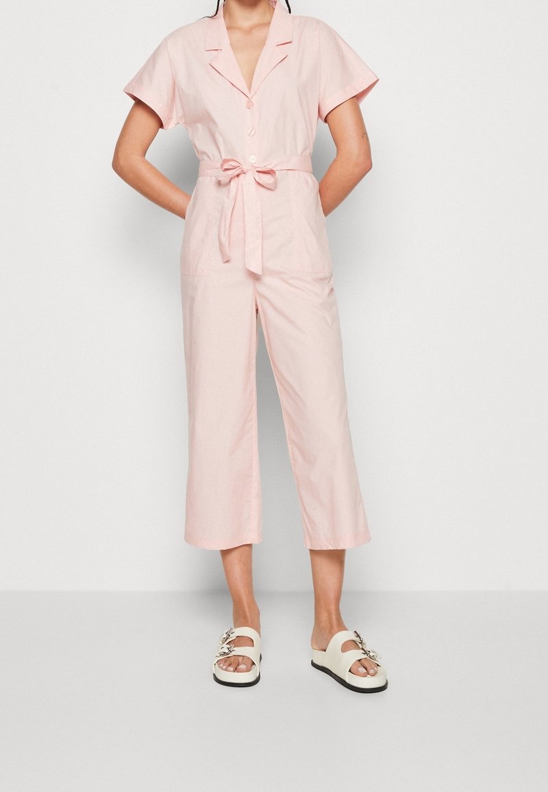 Light pink jumpsuit with short sleeves, tied waist, and cropped pants. Features a button-up front and two front pockets.