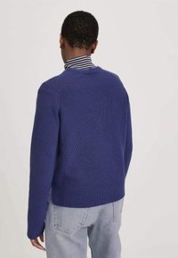 Mann, der sich von der Kamera abwendet, trägt einen blauen Strickpullover über einem schwarz-weißen gestreiften Rollkragenpullover und hellblauen Jeans vor einem schlichten Hintergrund.