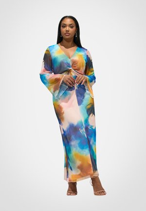 Femme portant une robe longue à motifs tourbillonnants multicolores avec des manches évasées, debout les mains légèrement jointes, fond neutre.