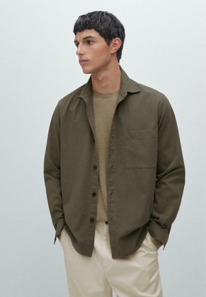 Olivgrön skjorta med knappar och en enda bröstficka, bärs över en beige t-shirt med rund halsringning, tillsammans med ljusa byxor.