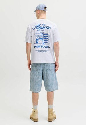 Homme portant une casquette bleu clair, un t-shirt blanc avec un imprimé boîte de sardines et le texte « Portugal », un short en jean et des bottes beiges, debout dos à la caméra.