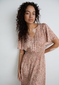 Vestido rosa con estampado, escote en forma de V, mangas cortas y con volantes, y cintura fruncida. La tela presenta manchas y remolinos en una textura suave.