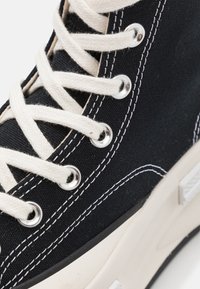 Black canvas sneaker with white stitching and laces, featuring metal eyelets, a rubber sole, and a textured upper.