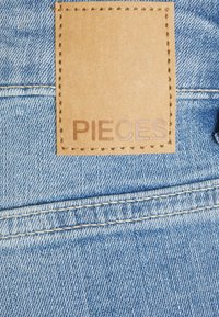 Um tecido de denim com uma tonalidade azul clara apresenta um retângulo de couro castanho cosido, exibindo o texto "PIECES" de forma proeminente.