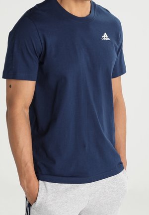 Homme portant un t-shirt Adidas bleu marine et un pantalon de jogging gris clair, mains dans les poches, debout devant un fond uni.