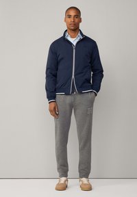 Marineblå bomberjakke med hvide detaljer, parret med grå sweatpants med et logo. Modellen bærer beige sneakers og en lyseblå skjorte under.