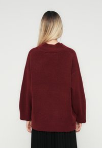 Kobieta z prostymi blond włosami, ubrana w luźny bordowy sweter i czarną plisowaną spódnicę, stojąca na tle jasnej jednolitej ściany.
