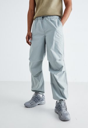 Person trägt hellgraue, locker sitzende Hose mit elastischem Bund und graue Sneakers, steht vor einem einfarbigen hellen Hintergrund.