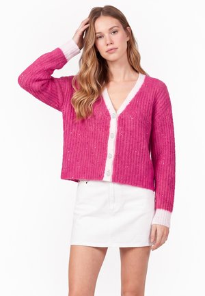 Junge Frau mit langen blonden Haaren, die eine leuchtend pinke geknöpfte Strickjacke und einen weißen Rock trägt, steht vor einem schlichten Hintergrund.