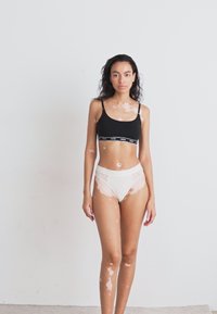Soutien-gorge de sport noir avec des bordures et un logo blancs, associé à des panties blancs avec des détails en dentelle. Le modèle se tient devant un fond clair.