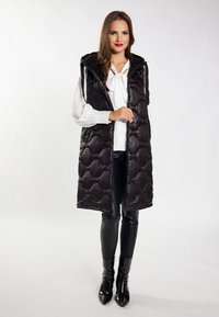 Gilet imbottito nero con cappuccio, caratterizzato da un design trapuntato, zip frontale e tasche laterali, indossato sopra una blusa bianca e pantaloni neri.
