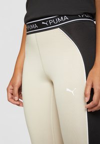 Puma tajice s dizajnom u crnoj i bež boji. Imaju crni pojas s bijelim logotipom i kontrastnim bočnim panelima. Glatka tekstura.