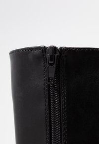 Gros plan sur une tige de botte en cuir noir et daim avec une fermeture éclair noire verticale et des coutures détaillées sur un fond clair.