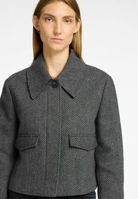 Giacca grigia con pattern a spina di pesce, colletto ampio, due tasche frontali e chiusura con bottone nero. Design strutturato e leggermente cropped.