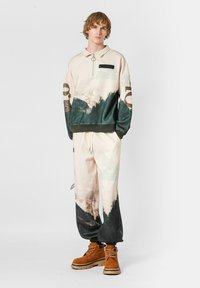 Suéter de patrones crema y verde con media cremallera y bolsillo en el pecho negro, combinado con pantalones a juego y botas marrones con acentos negros.