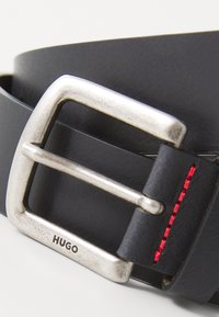 Cinturón de cuero negro con una hebilla rectangular plateada, que presenta un detalle de costura roja y "HUGO" grabado en la hebilla.